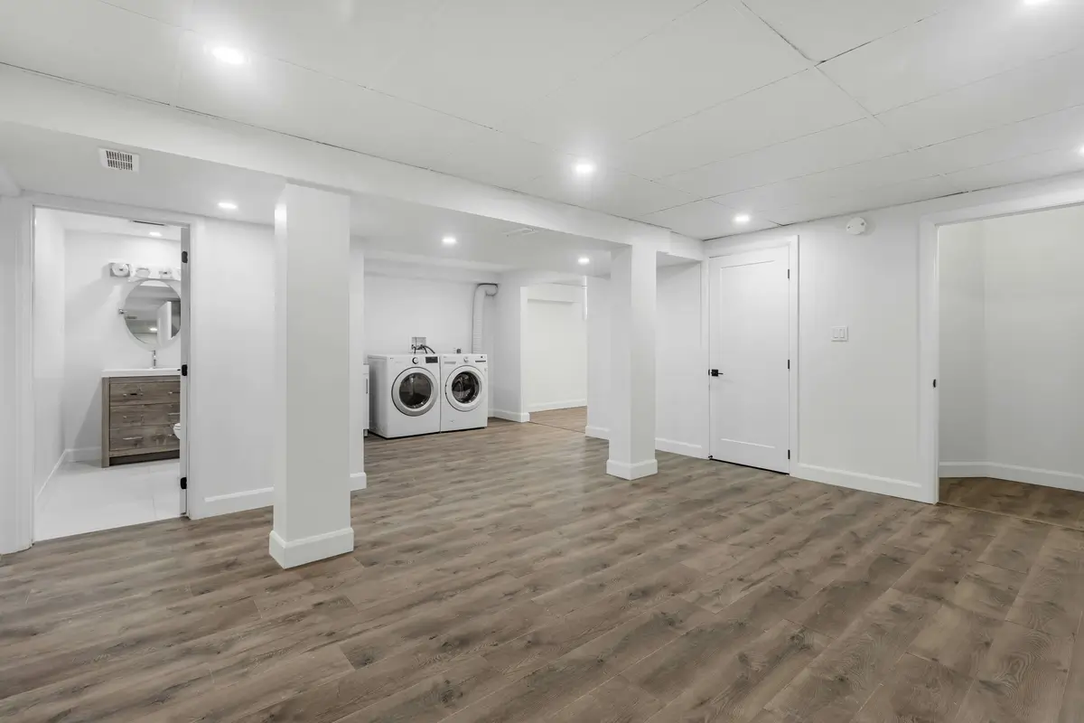 Modern finished basement with hardwood flooring, bright recessed lighting, and a dedicated laundry area featuring a washer and dryer. The space includes a bathroom and additional storage rooms, showcasing a functional and stylish design. Maximize your home's value and space by finishing your basement today!