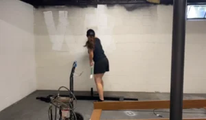 A woman painting a white concrete wall in a basement with tools and equipment in the foreground. Transform your basement today with Newton Basement Finishing—specialists in creating cozy, functional spaces.