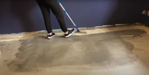 A person using a floor mop to clean and prepare a basement concrete floor for painting, ensuring smooth and even application.