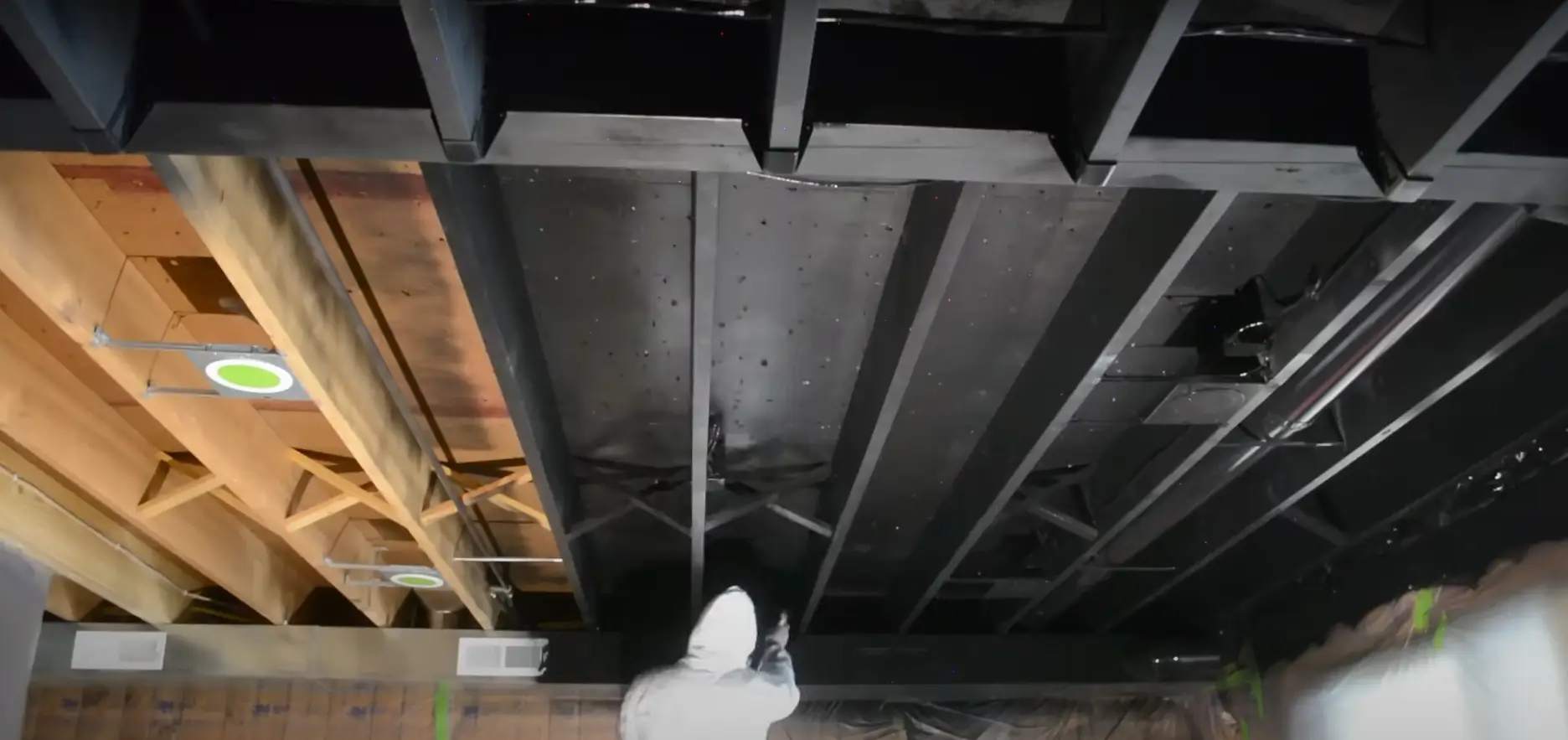 A person in protective gear sprays black paint on an exposed wooden and metal basement ceiling, creating a sleek, modern look.