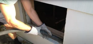 A person wearing work gloves installs a glass block window in a basement, using a vacuum to clean the opening before securing the new window.